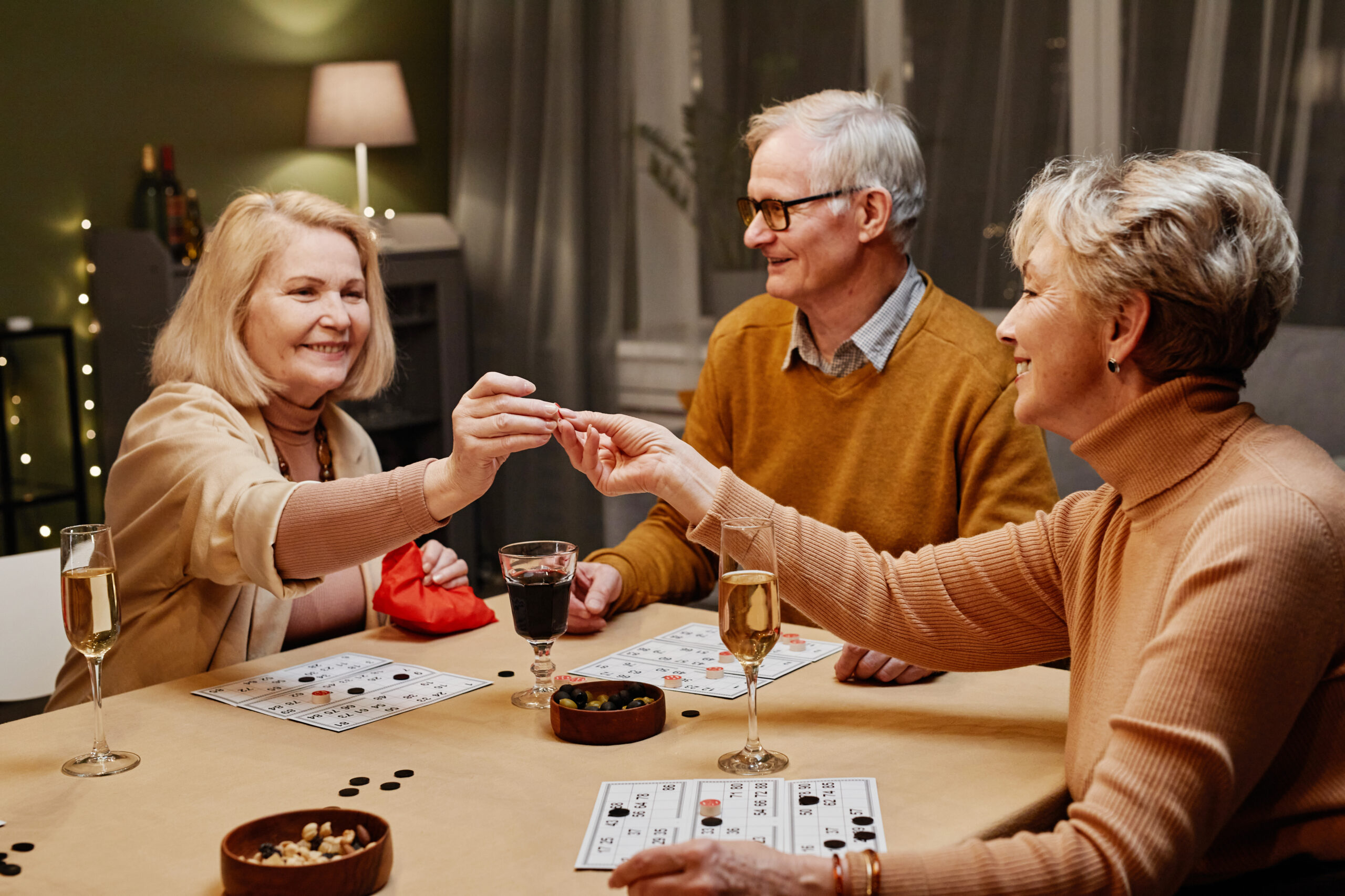 Two aged Caucasian women and man sitting at wooden table with snack and drinks during party and playing board game