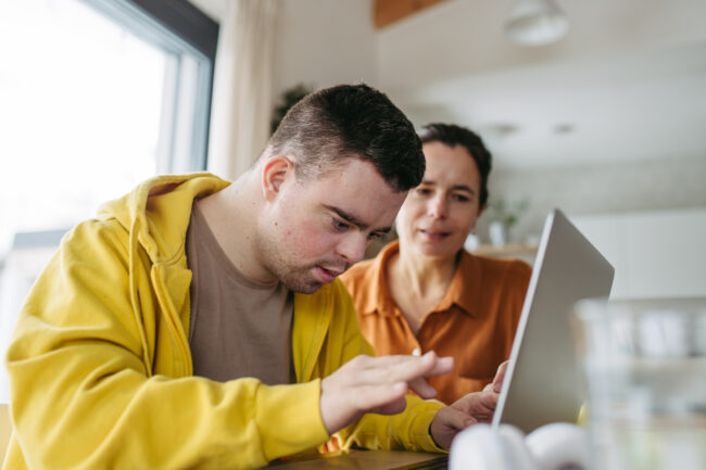 Mom teaching young man with down syndrome, using laptop. Telehealth consultation with doctor.