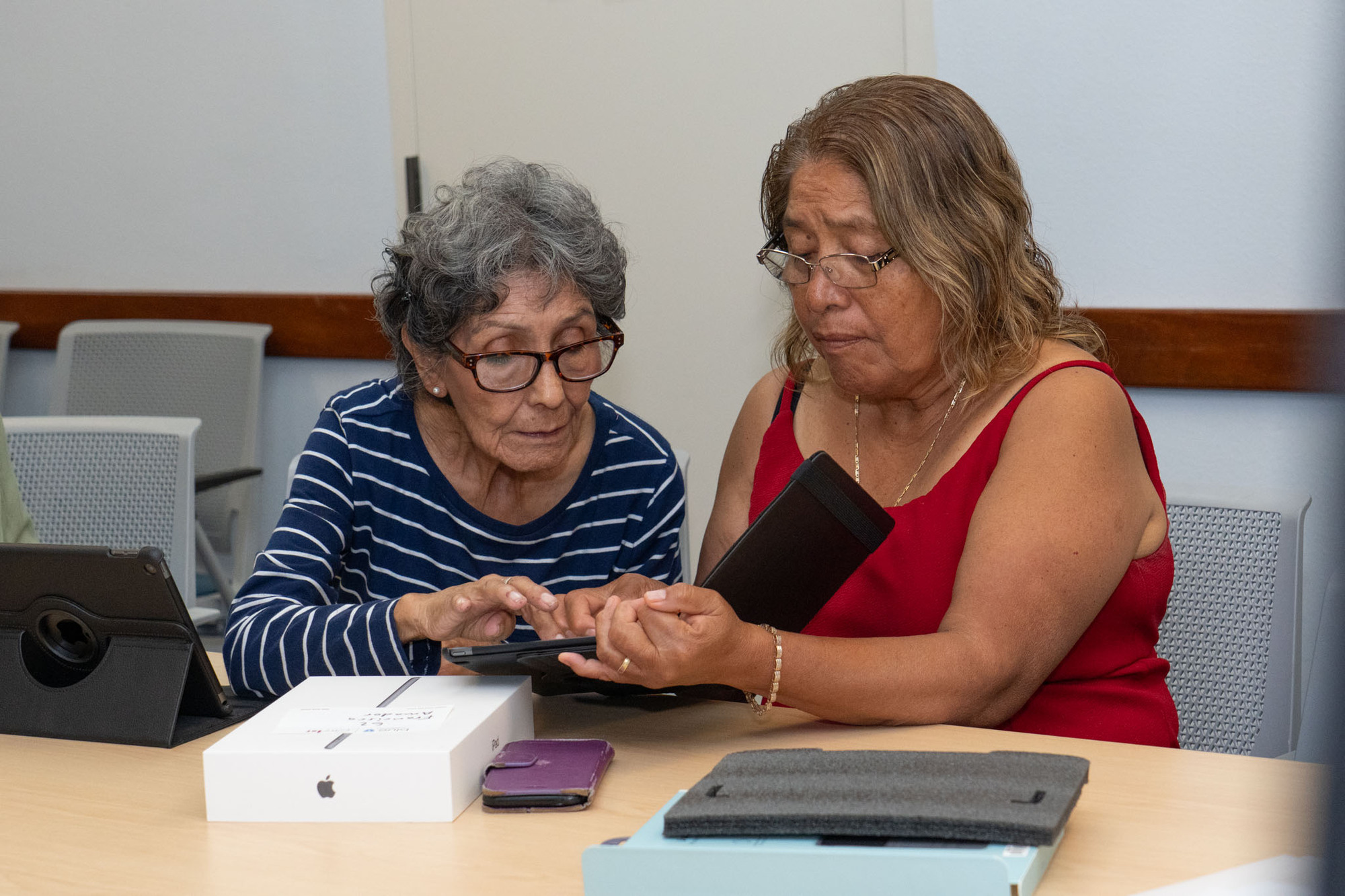 A middle aged woman with dark blonde hair, holds an iPad; an older woman with short grey hair touches its screen.