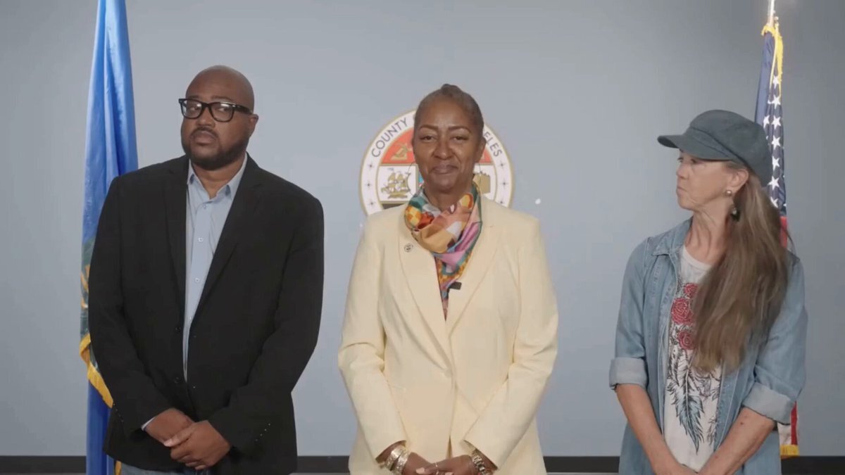 An African-American middle-aged woman in a cream suit smiles to the camera--with an African-American man on her left side; on her right, a brunette woman with long hair with a side ponytail--wearing a light blue baseball cap --looks toward her.