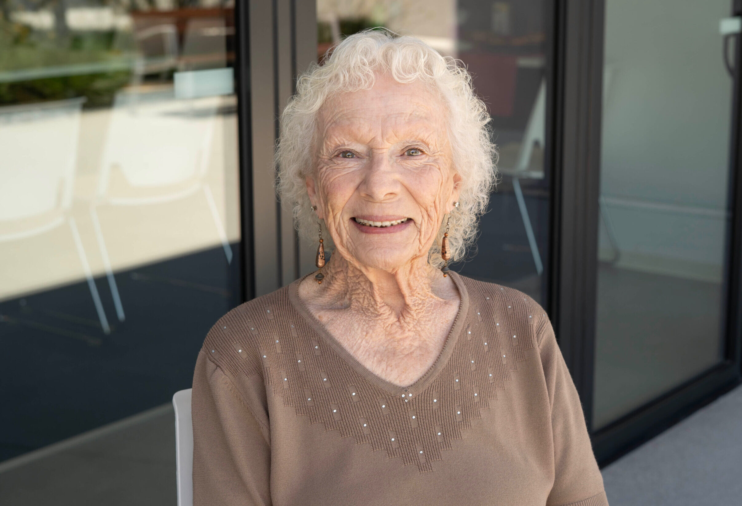 Margaret “Peggie” Reyna smiles outdoors, wearing a brown sweater with beaded details and earrings.
