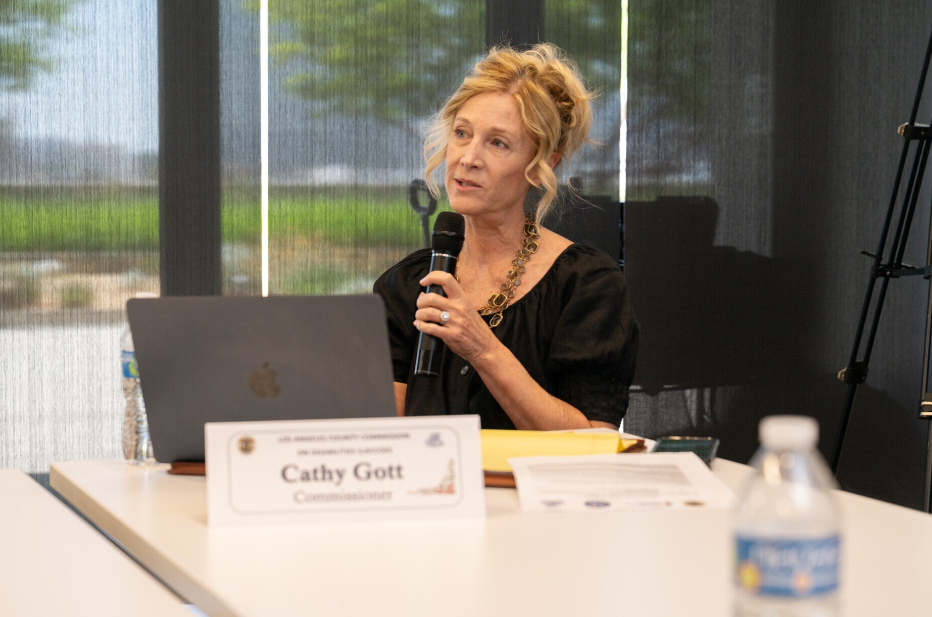 A blonde woman seated at a meeting table holds a microphone while speaking. A nameplate reading “Cathy Gott, Commissioner” sits in front of a laptop, along with papers, a folder, and a water bottle. Sunlight filters through tall windows with partially drawn shades, and greenery is visible outside.