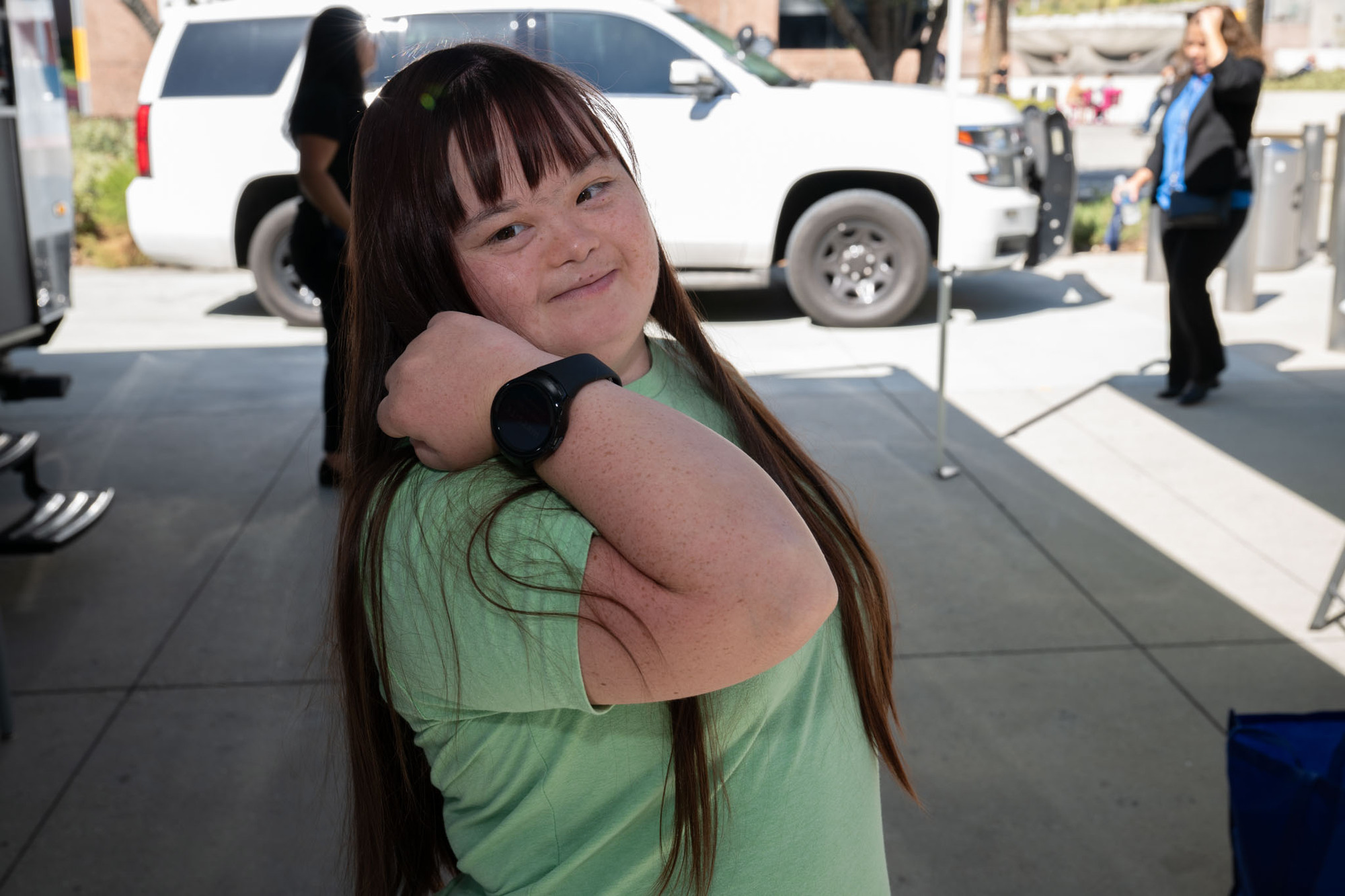 A young woman with long brown hair, born with Down Syndrome, smiles while showing off a black smartwatch on her wrist. She is standing outdoors in front of a white SUV and wearing a light green shirt.