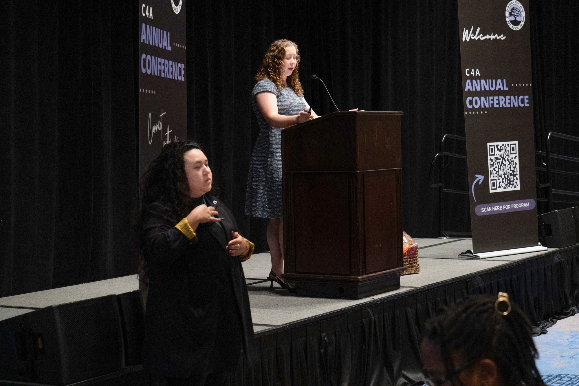 A young woman on the stage at the podium talking at the C4A Annual Conference. There is another lady to the bottom left conducting sign language for the audience.