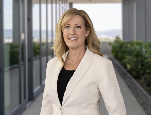 A professionally dressed woman in a beige blazer smiling in a modern office corridor.