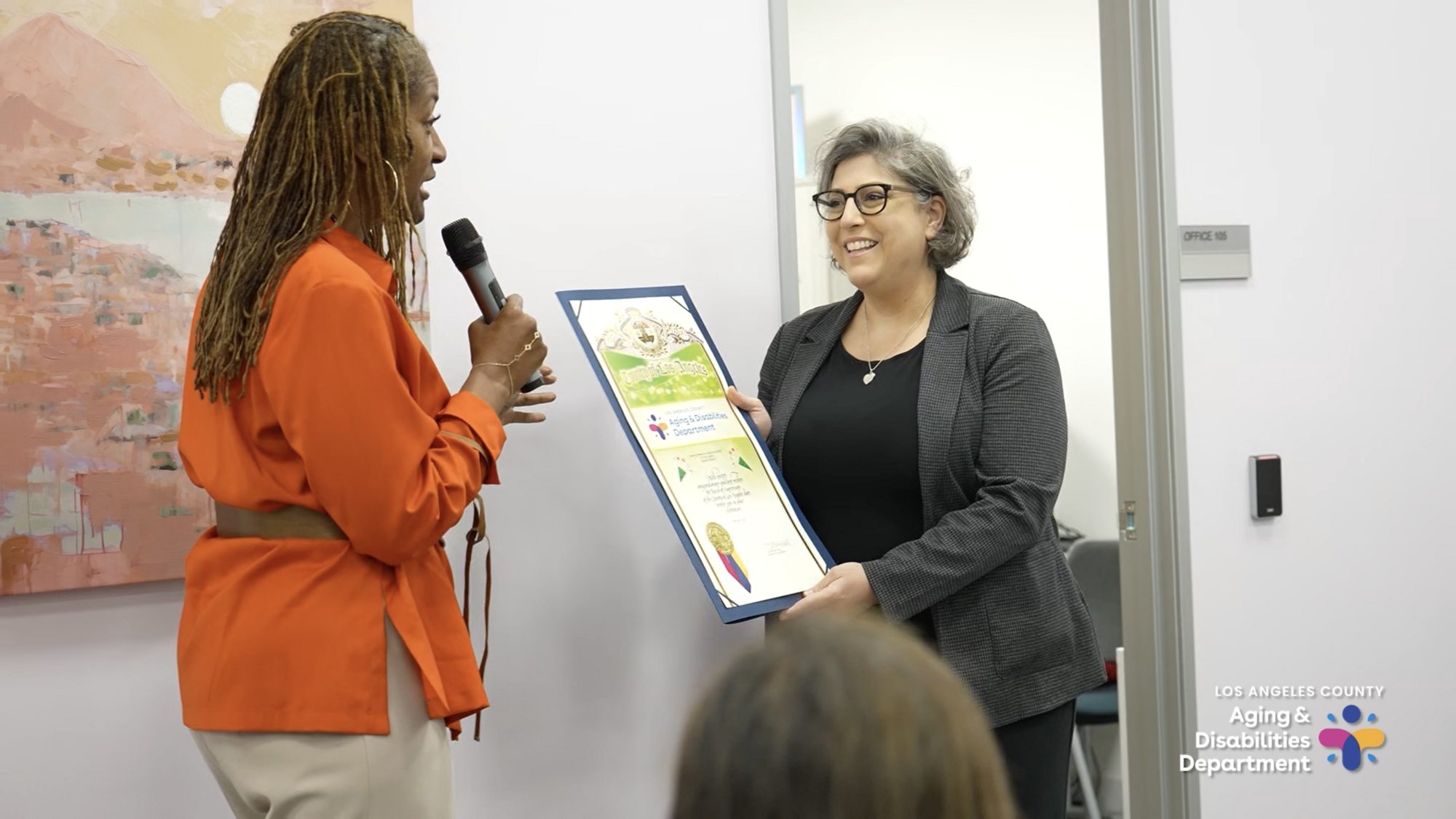 A woman with brown braids wearing an orange shirt holds up a microphone while another woman with gray hair holds up a poster.