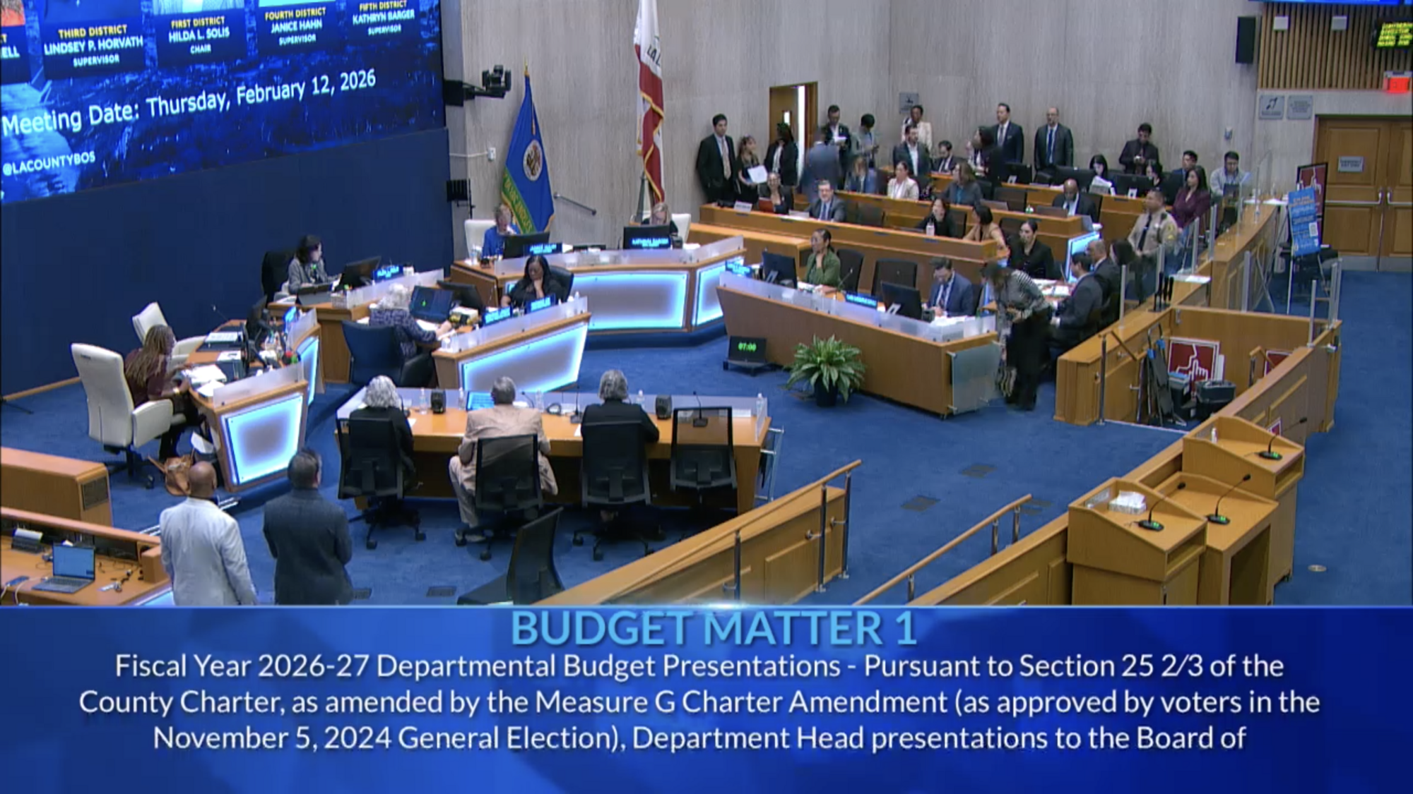 Several people sit in the LA County Board of Supervisors meeting chamber looking at a presentation.