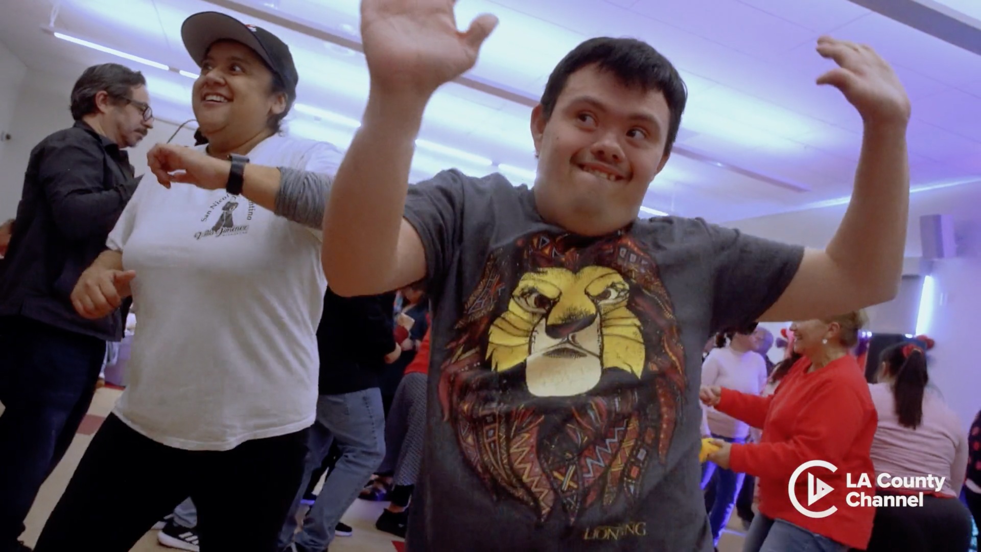 Several people are dancing at a community center. One man is in the front with a lion shirt and his hands up.