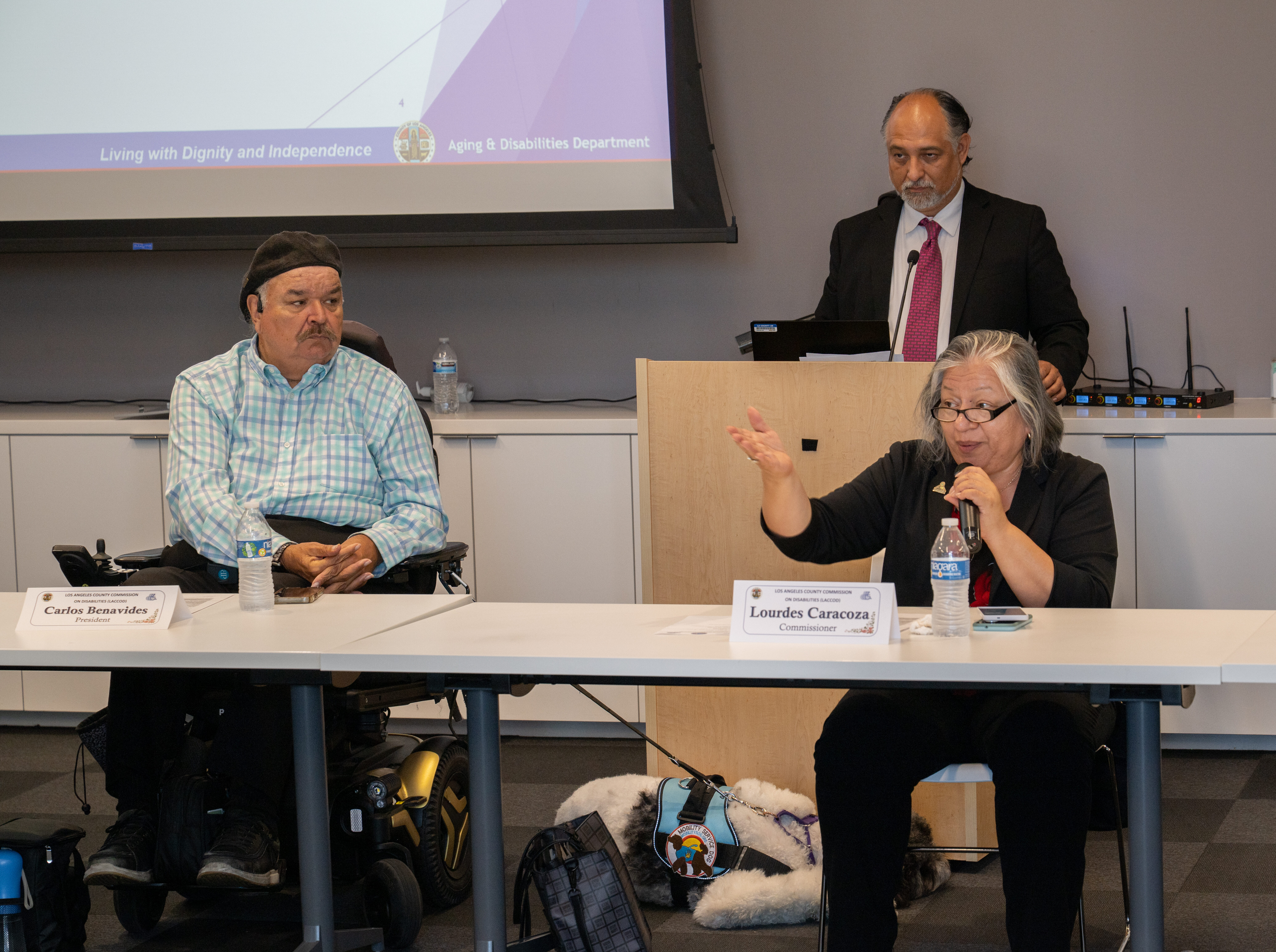 A man in a beret sits in a mobility device at a table next to an older woman speaking into a mic. A man stands at a podium behind the two people at the table.