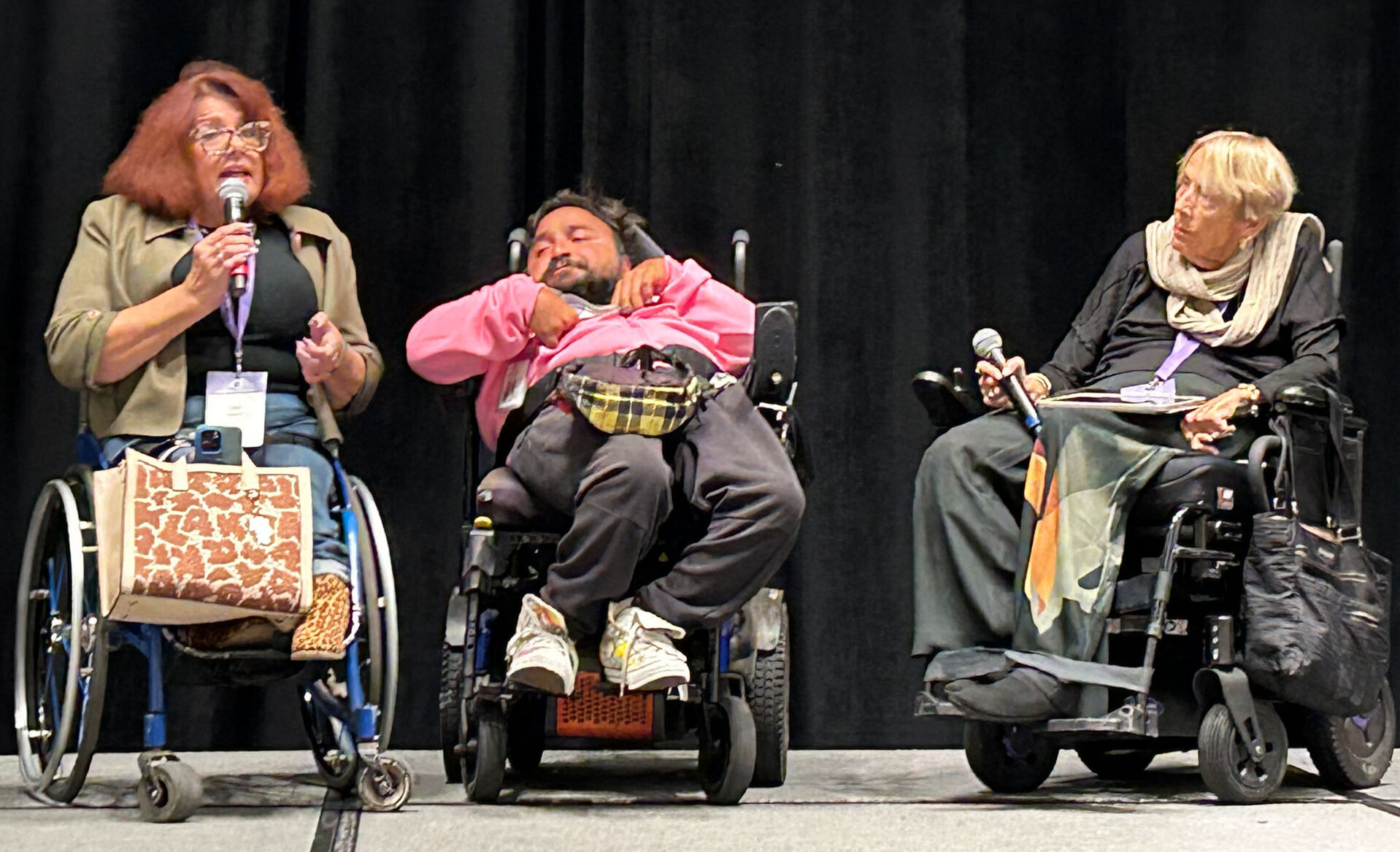 Three people sit in mobility devices on a stage during the C4A conference.