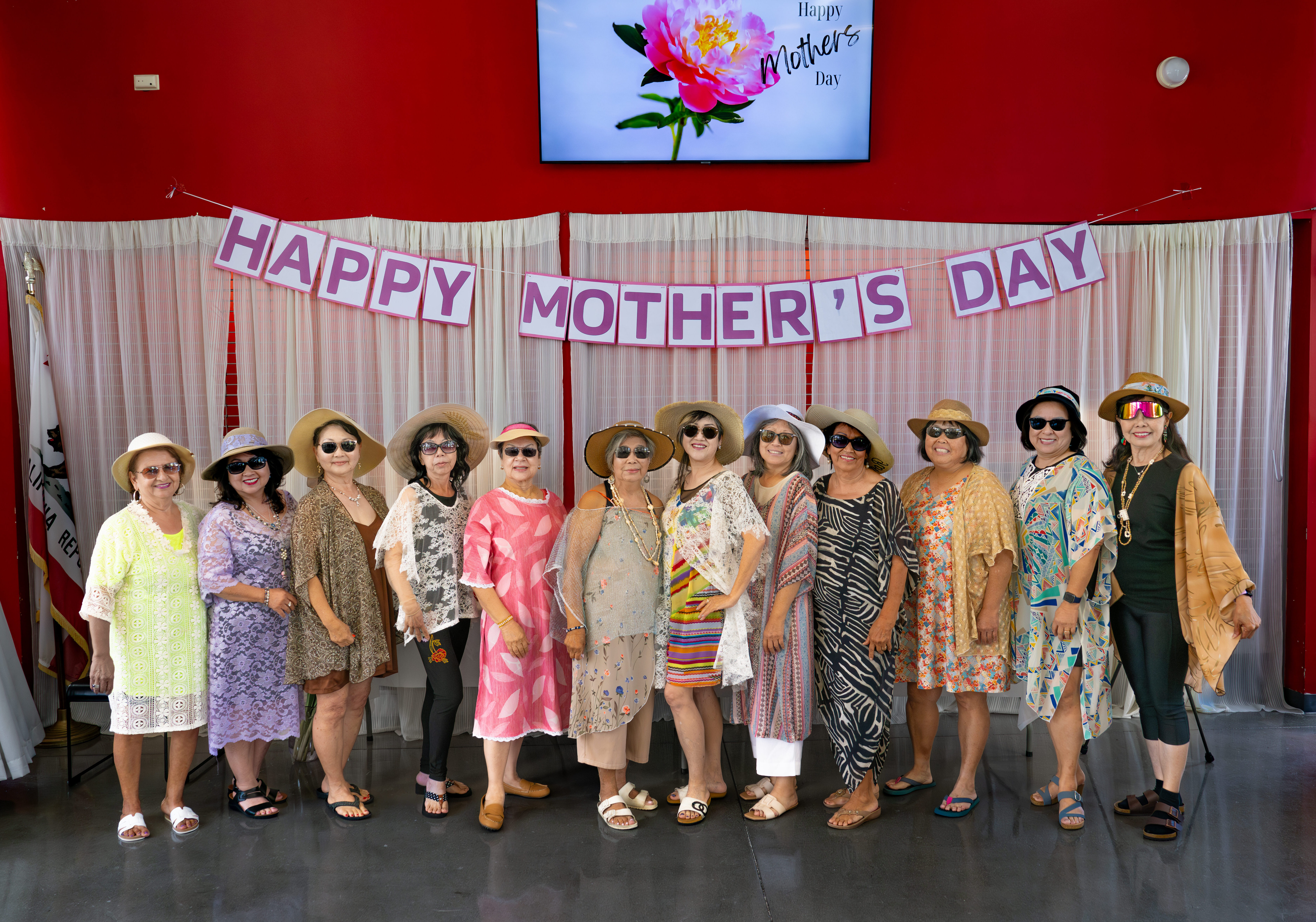A group of women stand together modeling clothes made in a sewing class in front of a banner that says "Happy Mothers Day"