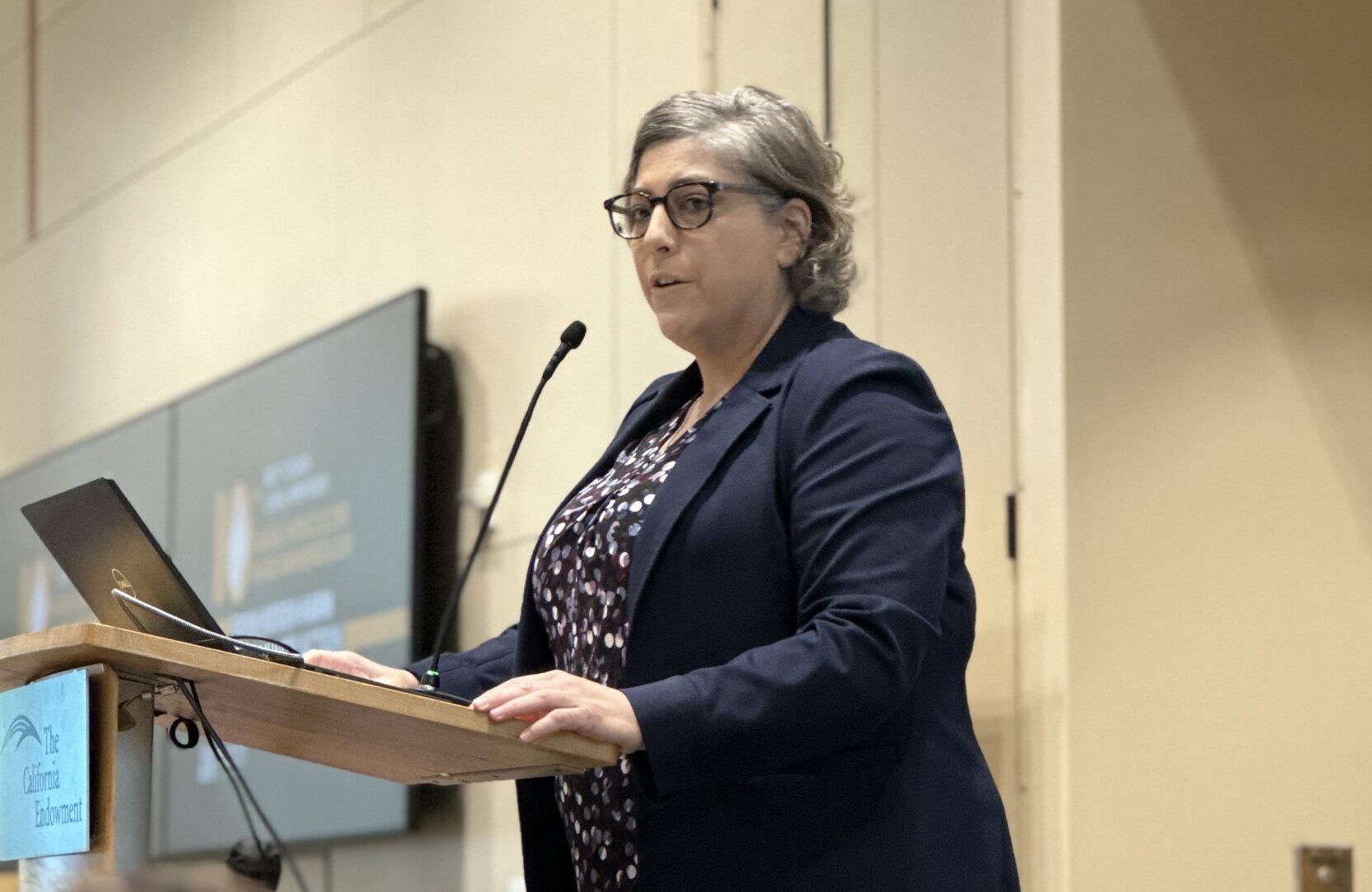 A woman stands at a podium, speaking at a conference.