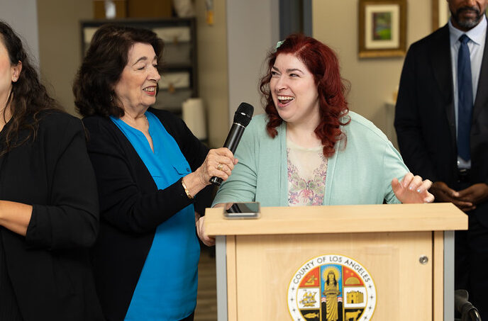 A woman stands at a podium while another woman holds up a microphone for her.